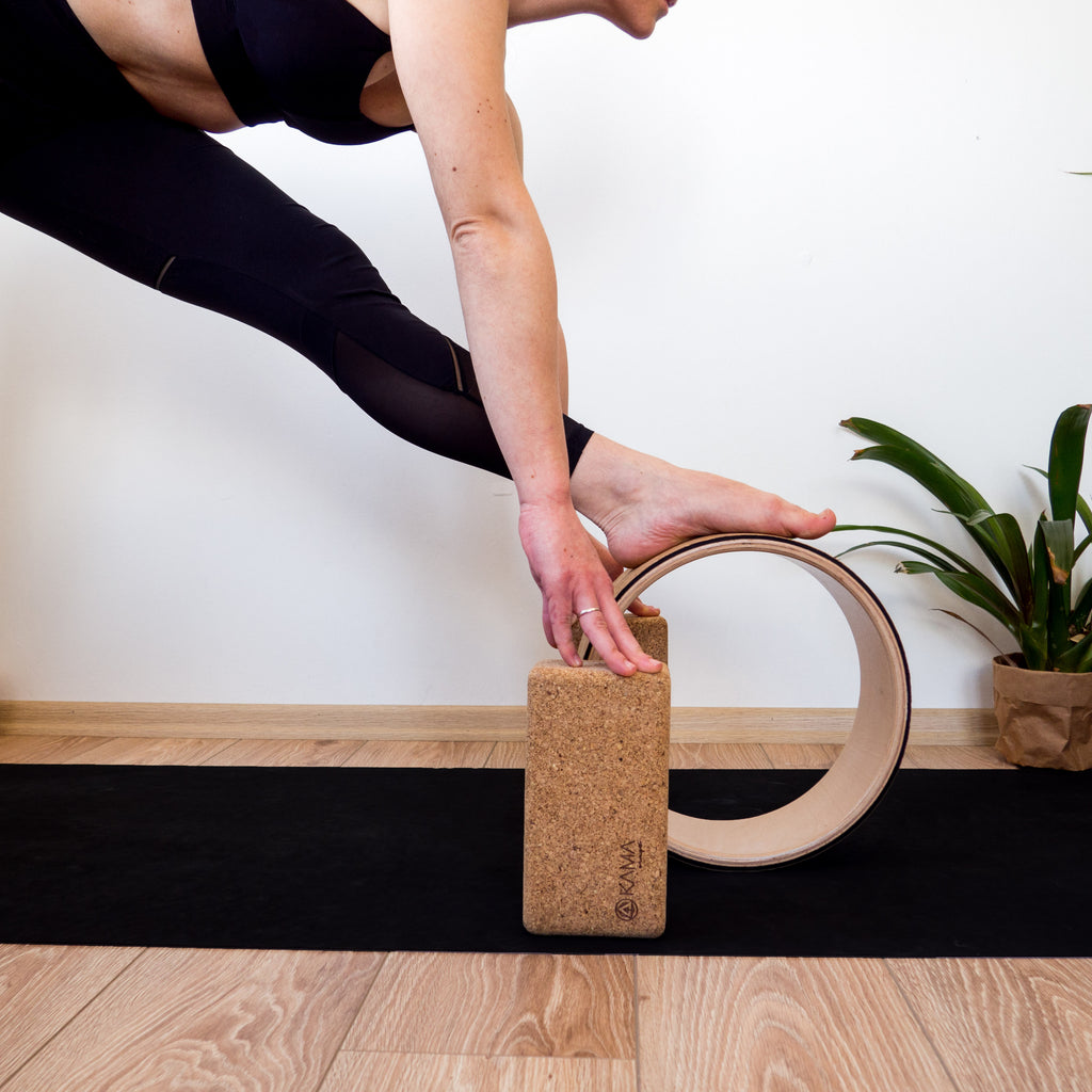Person using a cork yoga block and a yoga wheel on a wooden floor with a plant in the background